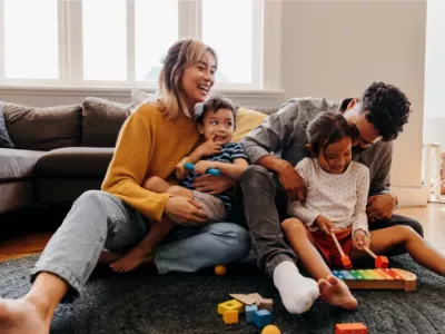 A young family sits on the floor, laughing and playing together with toys in a cosy living room.