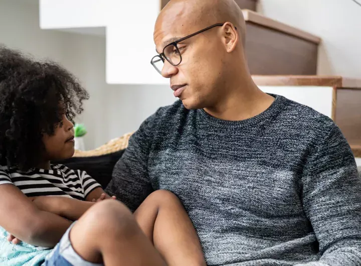 Child with arms folded sits on dad's lap on the sofa as they talk