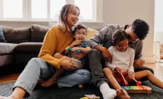 A young family sits on the floor, laughing and playing together with toys in a cosy living room.