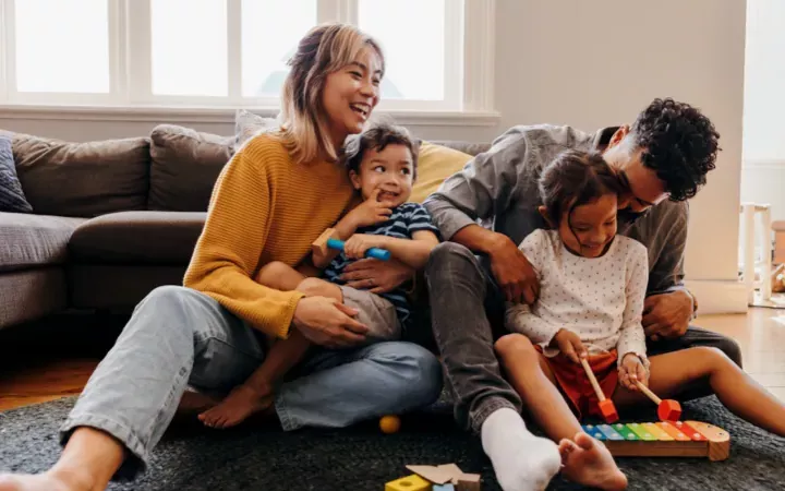 A young family sits on the floor, laughing and playing together with toys in a cosy living room.