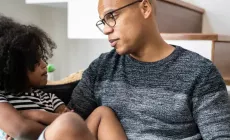 Child with arms folded sits on dad's lap on the sofa as they talk